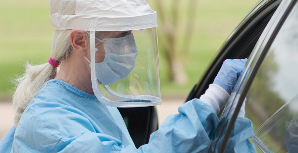 A public health worker wearing a medical face shield testing a motorist for COVID-19